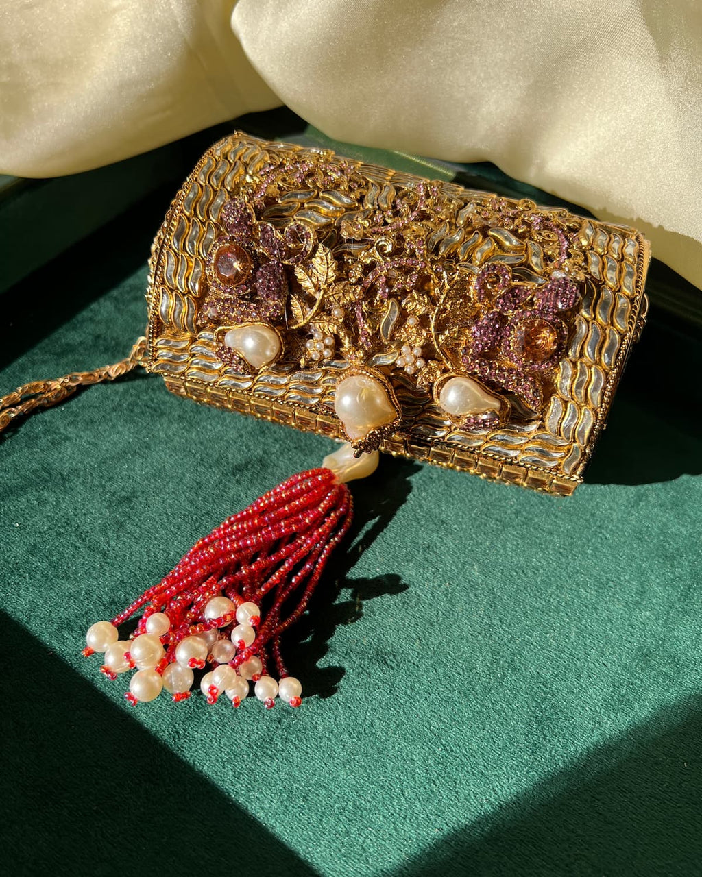 An ornate gold embroidered clutch bag with purple floral embellishments, faux pearls, and a red beaded tassel, displayed on a dark green velvet surface with cream fabric in the background.