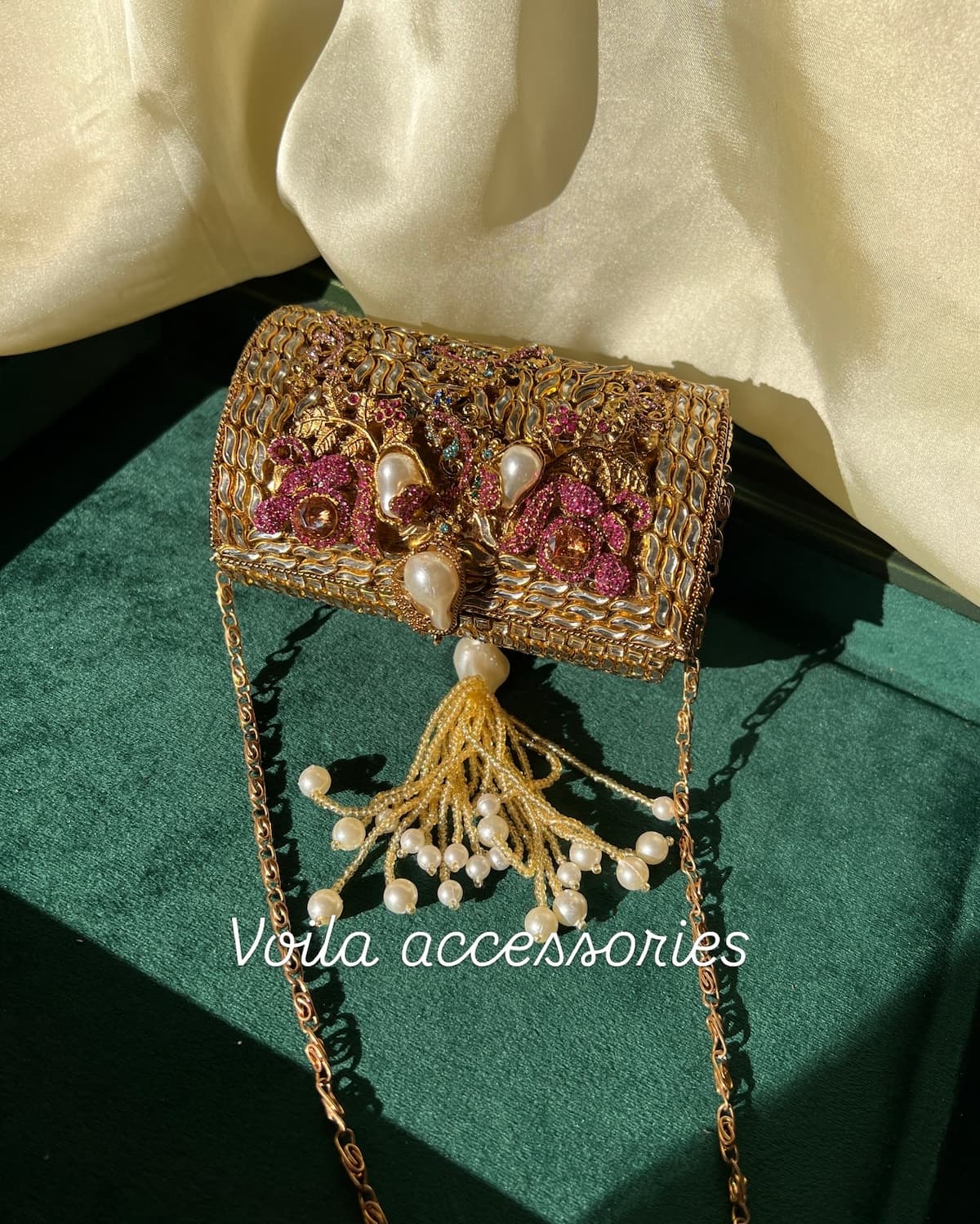 An ornate gold-toned embroidered clutch bag with pink floral embellishments, faux pearls, and a tassel, displayed on a dark green velvet surface with cream satin fabric in the background.