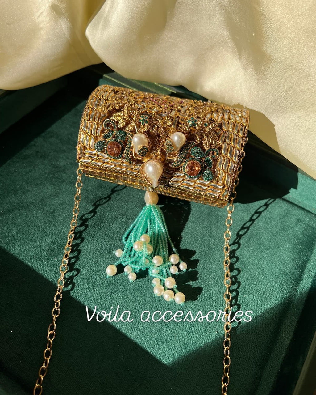 An ornate gold clutch purse with intricate beadwork, faux pearls, and a turquoise tassel, displayed on a dark green velvet surface with cream fabric in the background. The text 'Voila accessories' is overlaid at the bottom.