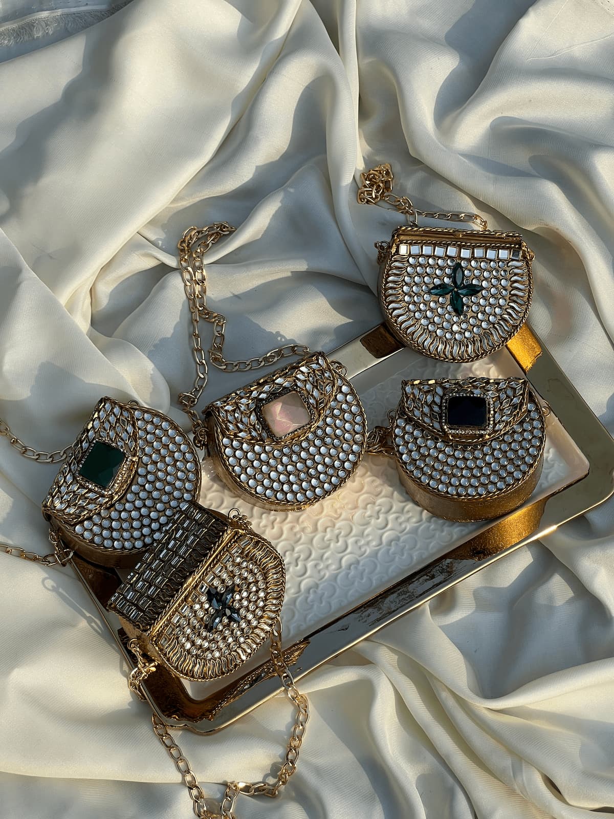 Four gold-toned embellished handbags arranged on a gold tray over draped white satin fabric.
