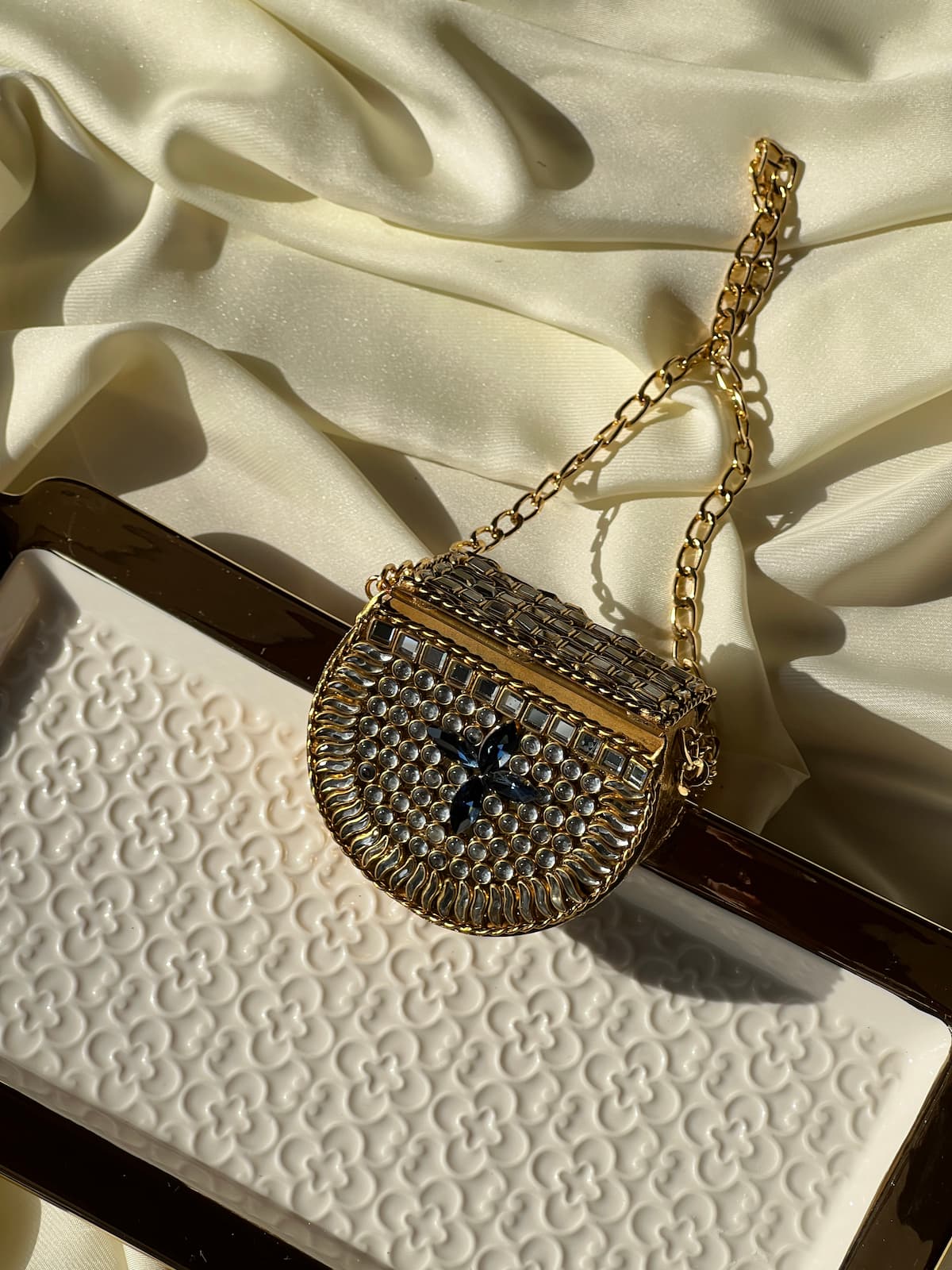 A small gold chain shoulder bag with a textured, rhinestone-embellished body and a dark blue cross detail on the front, displayed on a white patterned surface with draped cream satin fabric.