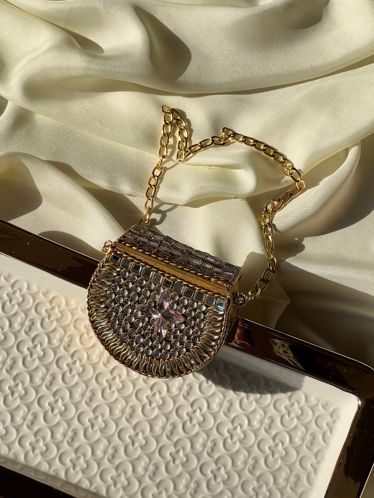 A gold-tone chain handbag with a textured rhinestone-embellished body and a pink crystal flower detail, displayed on draped cream satin fabric.