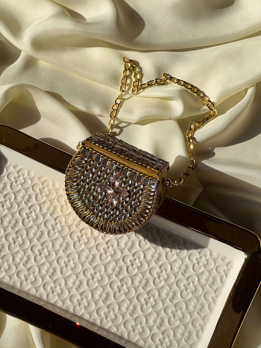 A small structured handbag with a gold-tone chain strap, covered in rhinestones and featuring a central pink crystal flower, displayed on a textured white surface with draped cream satin fabric.