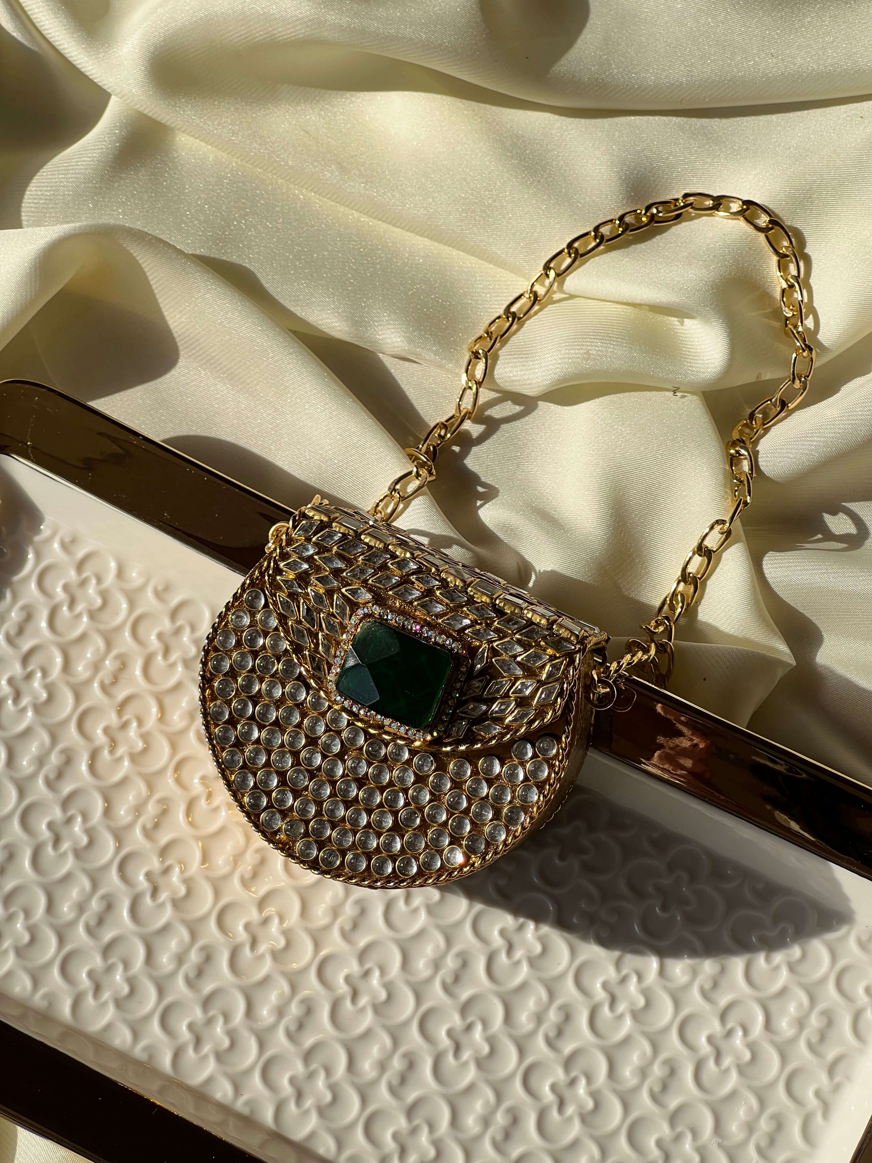 A gold chain handbag with a rhinestone-embellished body and a central dark green rectangular stone, styled on a cream satin fabric and a white textured clutch.