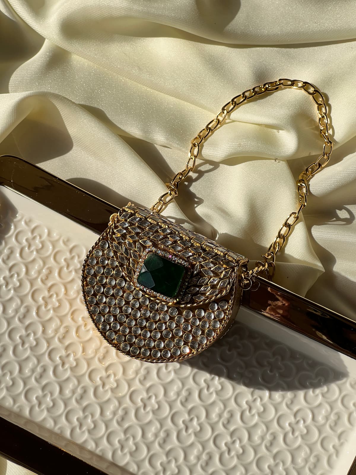 A gold chain handbag covered in clear rhinestones, featuring a green faceted crystal accent on the front flap, displayed on a cream-colored textured surface with draped satin fabric in the background.