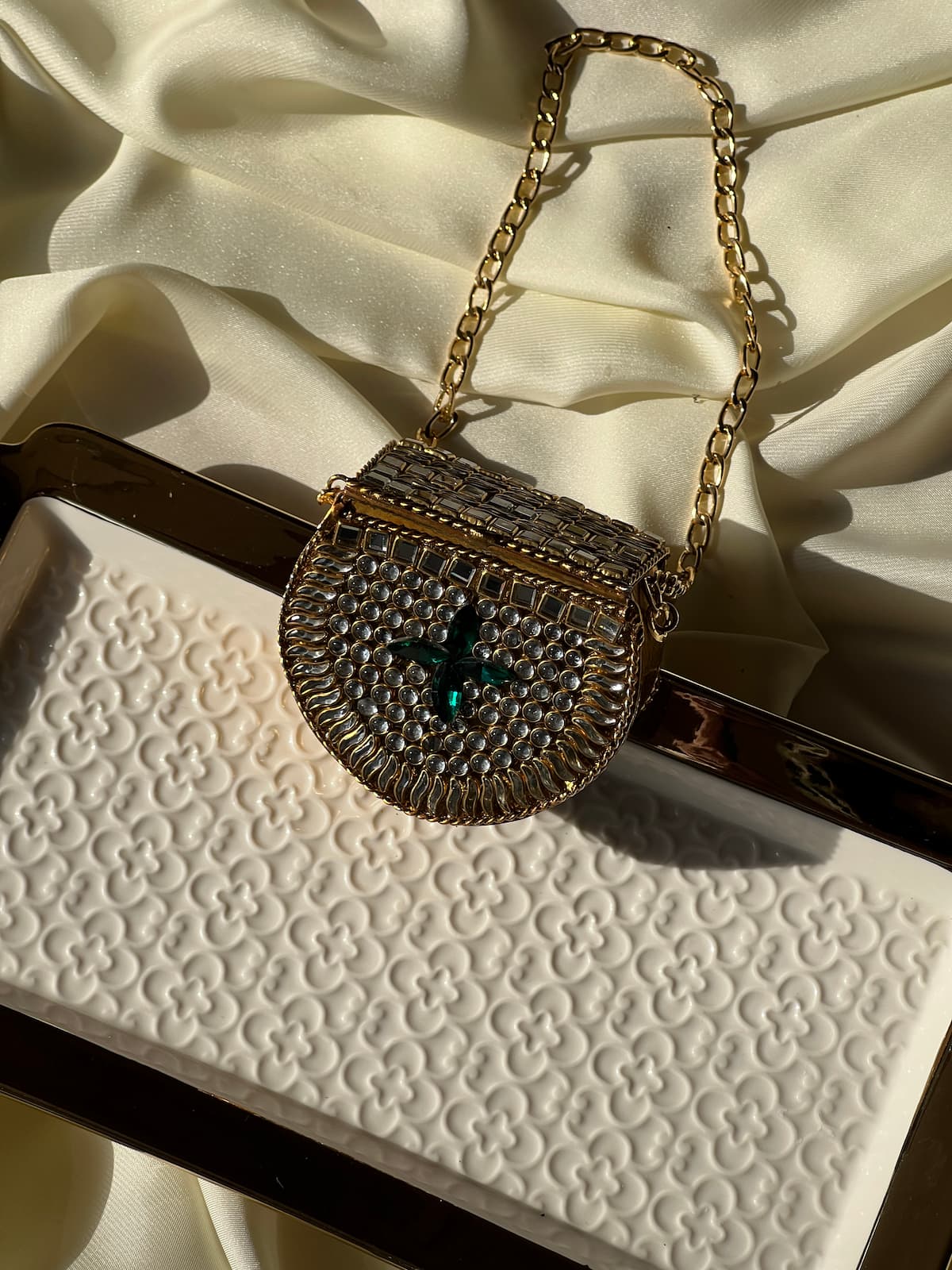 A small structured evening bag with a gold-tone chain strap, covered in silver rhinestones, featuring a central green gemstone, resting on a white embossed clutch and cream satin fabric.