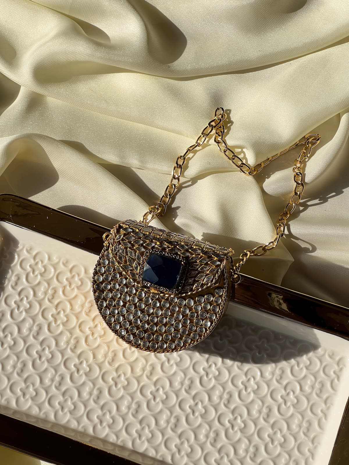 A gold chain handbag with a rhinestone-encrusted body and a dark blue square accent, styled on a cream satin fabric and a textured cream clutch.