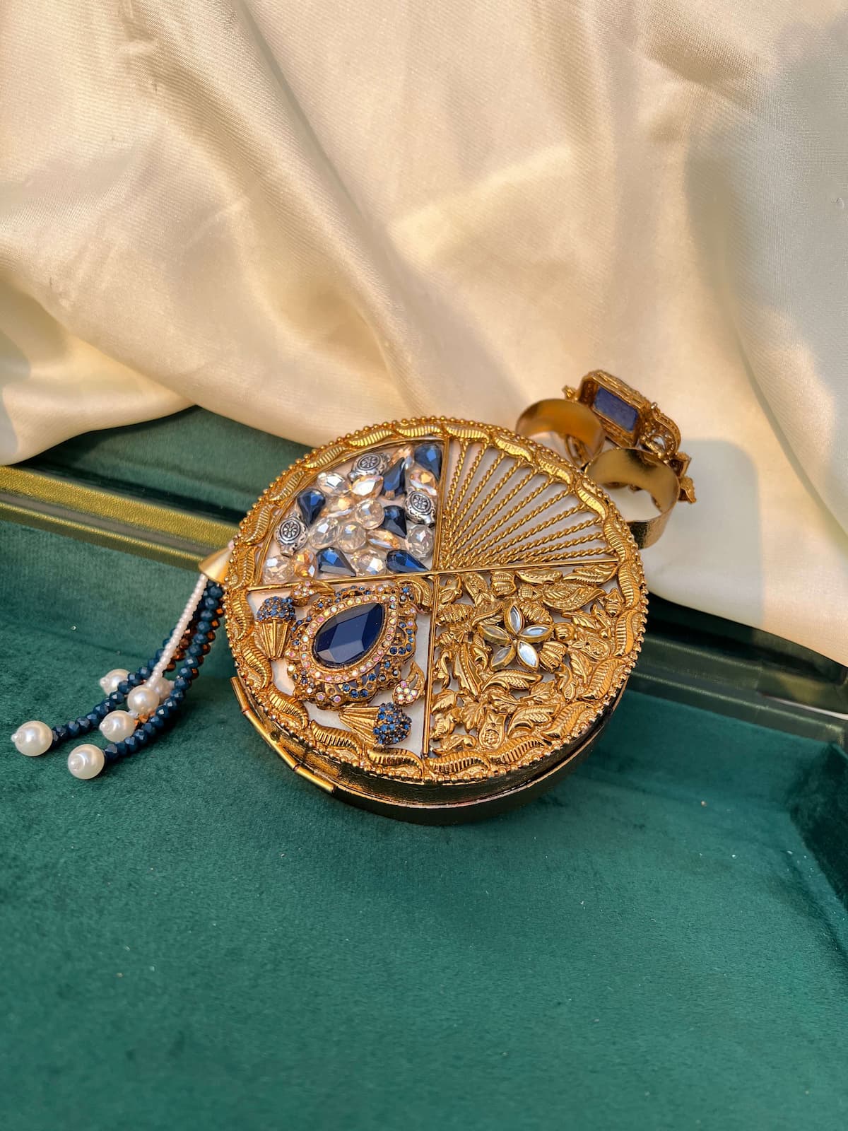 An ornate round gold clutch purse with intricate filigree detailing, set against a draped cream satin fabric and a dark green velvet surface. The clutch features a four-quadrant pattern of blue sapphires, white pearls, and clear crystals, with a blue and gold beaded tassel hanging from the side.