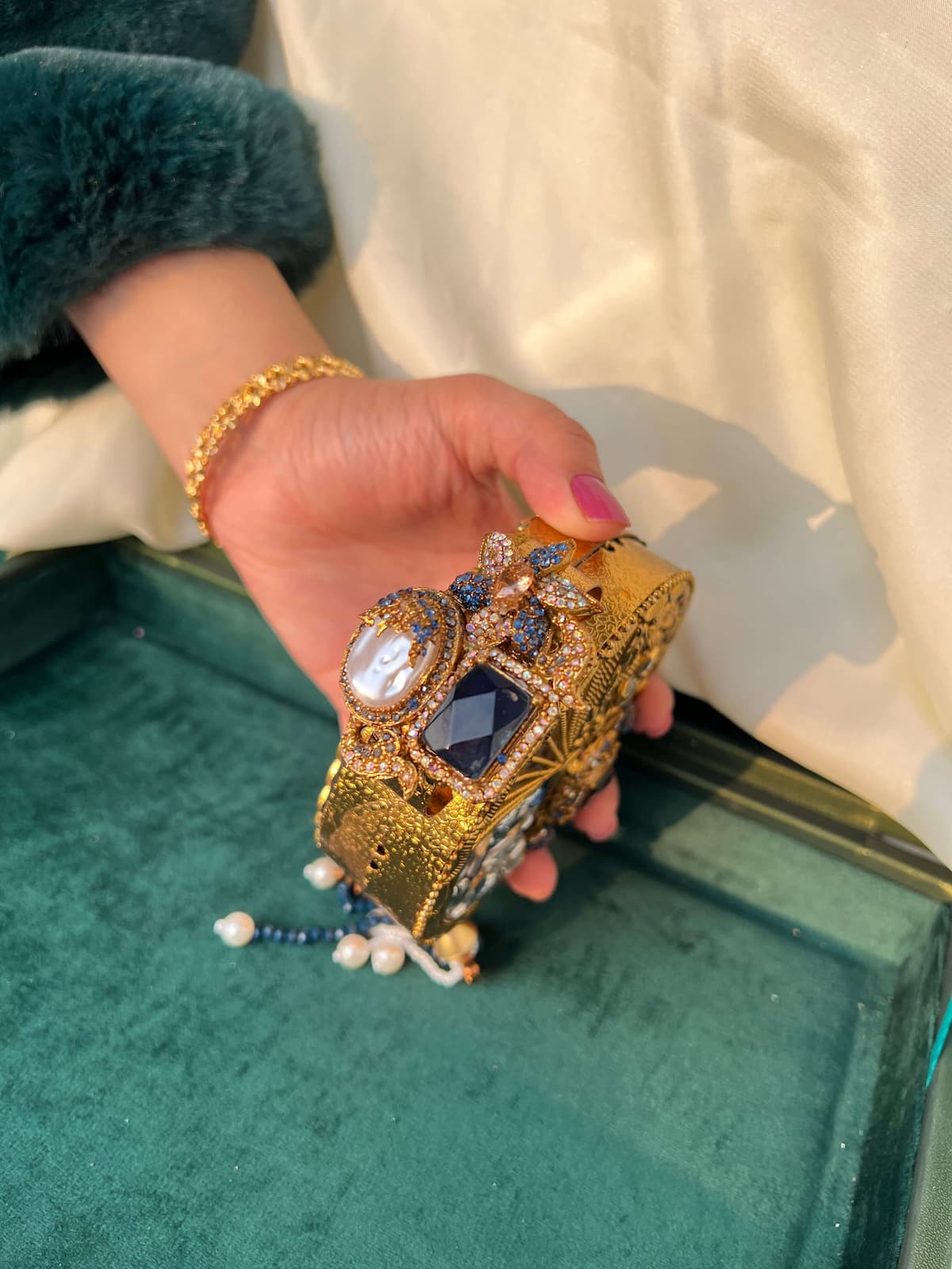 A hand holding an ornate gold clutch bag decorated with crystals, blue gemstones, and pearl-like bead tassels, set against a background of cream fabric and a green velvet tray.