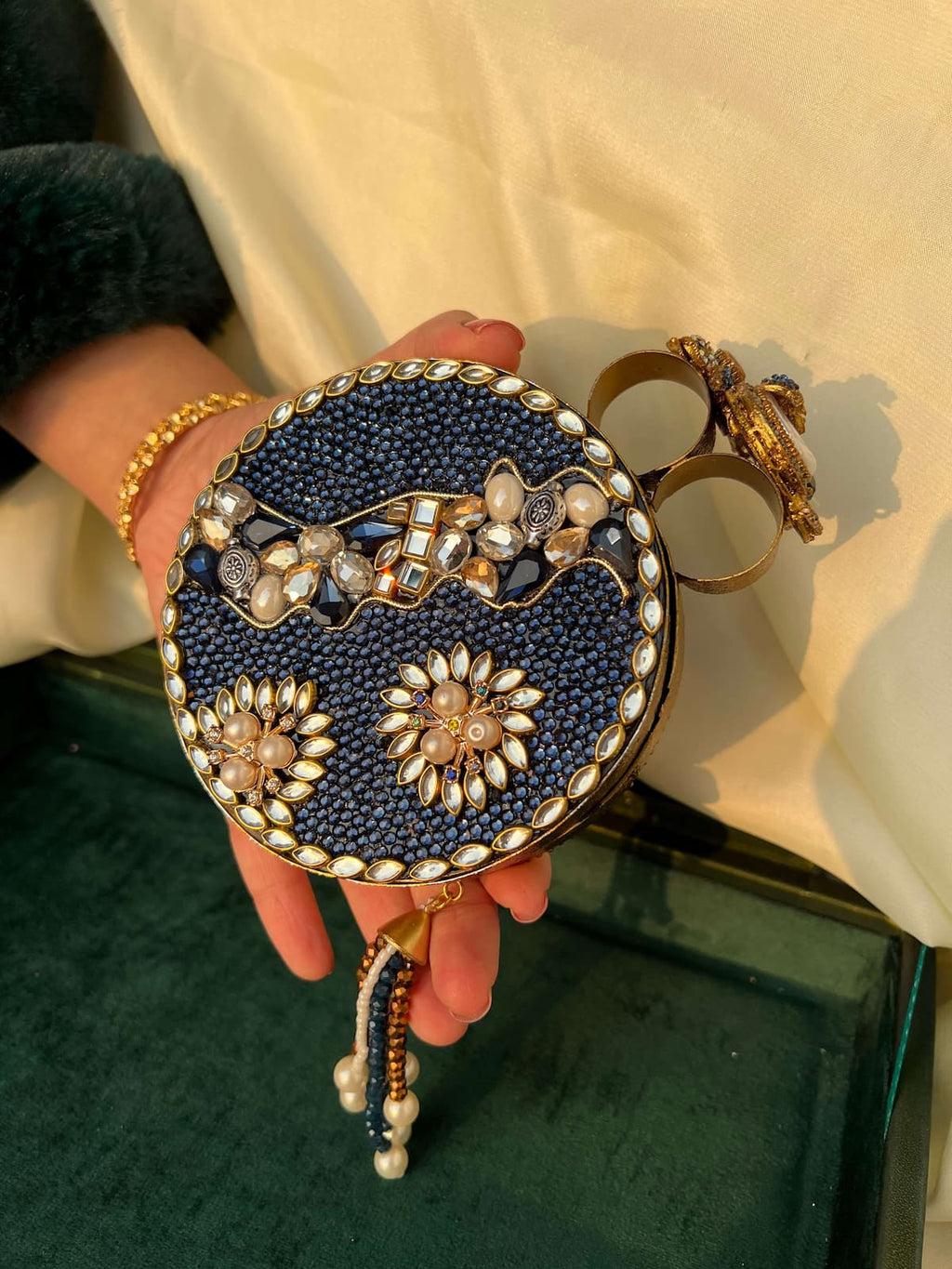 A hand holding a round navy blue beaded clutch bag with gold-tone metal trim, pearl-like embellishments, and a decorative clasp, set against a cream fabric background.